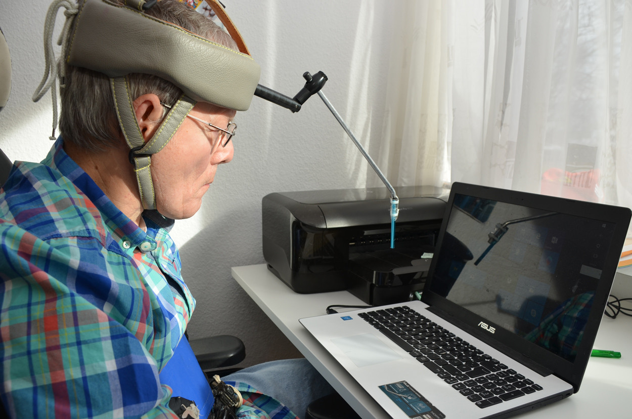 Ein Mann sitzt vor einem Laptop und tippt mit einem Stift, der an einem Gestellt auf seinem Kopf befestigt ist, auf die Tastatur.