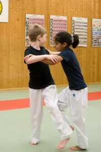 Ein blonder Junge und ein dunkelhaariges Mädchen tragen Sportbekleidung und kämpfen gegeneinander in einer Sporthalle.