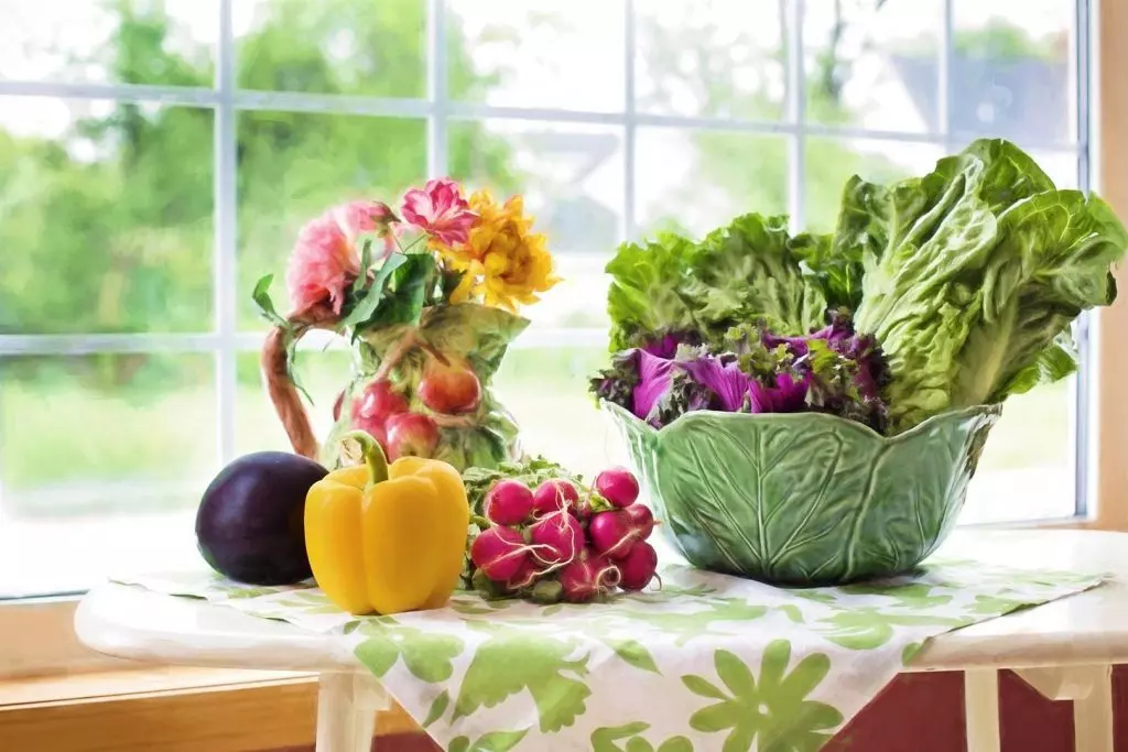 Auf einem Tisch, der vor einem Fenster mit Blick ins Grüne steht, liegen eine Aubergine, eine gelbe Paprika, rote Radieschen und eine grüne Schale mit Salat. Außerdem steht noch eine Vase mit bunten Blumen auf dem Tisch.