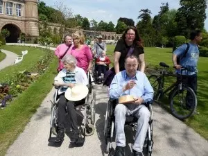 Auf dem Bild sind ein Mann und eine Frau zu sehen, die im Rollstuhl sitzen und von zwei Frauen durch den Botanischen Garten in Karlsruhe geschoben werden.