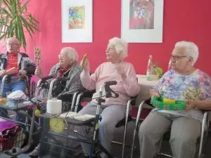 Auf dem Bild sind drei Seniorinnen zu sehen, die auf Stühlen sitzen und in die Hände klatschen.