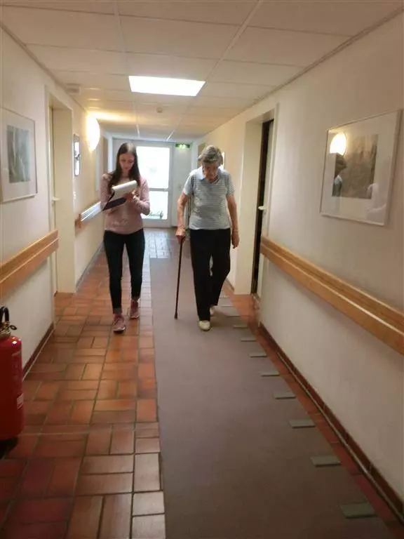 Eine junge Frau macht sich Notizen und läuft mit einer Seniorin in einem Flur einen Gang entlang.
