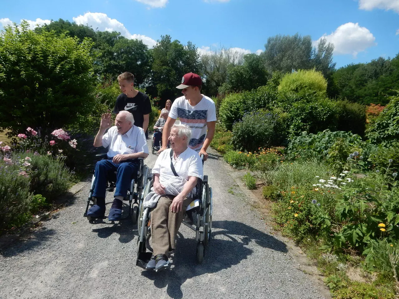 Zwei Senioren werden von zwei jungen Männern mit dem Rollstuhl durch eine blühende Landschaft geschoben.