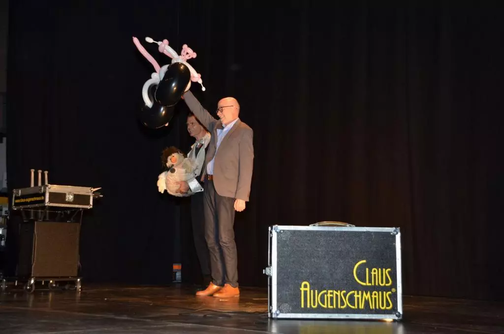 Ein Mann steht auf einer Bühne und hält einen rosaroten Panther aus Luftballons in die Luft.