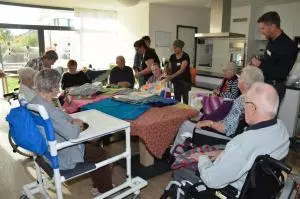 Eine Gruppe von Senioren und ihren Helfern sitzen an einem Tisch und schauen sich bunte Wolldecken an.