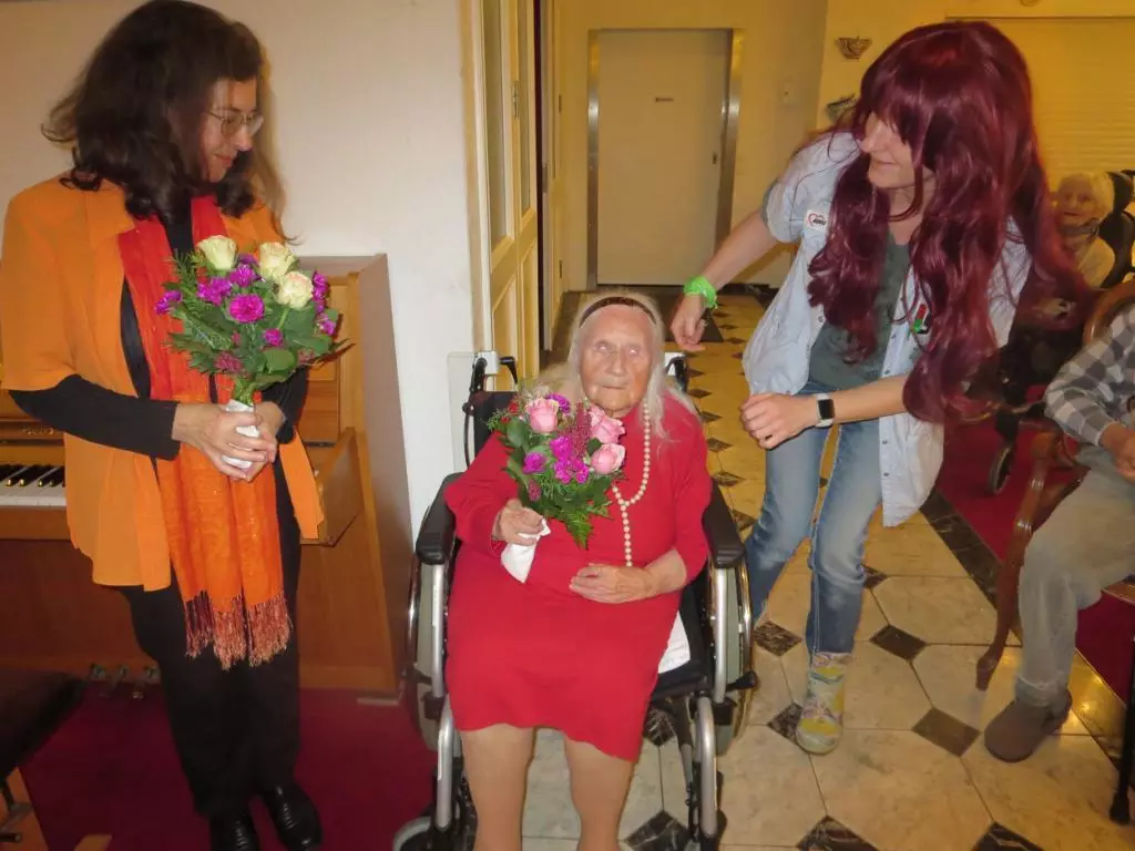 Eine Seniorin sitzt im Rollstuh, neben ihr stehen zwei Frauen mit Blumen in den Händen.