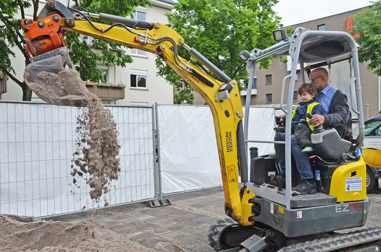 Ein Junge sitzt auf dem Schoß von einem Baggerfahrer und bedient die Maschine.