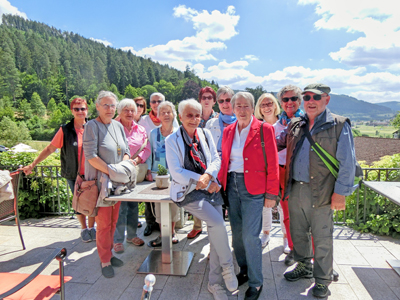Eine Gruppe von Menschen steht auf einer Dachterrasse im Hintergrund blauer Himmel und Berge.