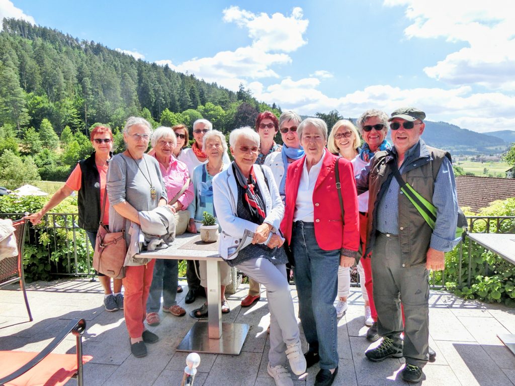 Eine Gruppe von Menschen steht auf einer Dachterrasse im Hintergrund blauer Himmel und Berge.