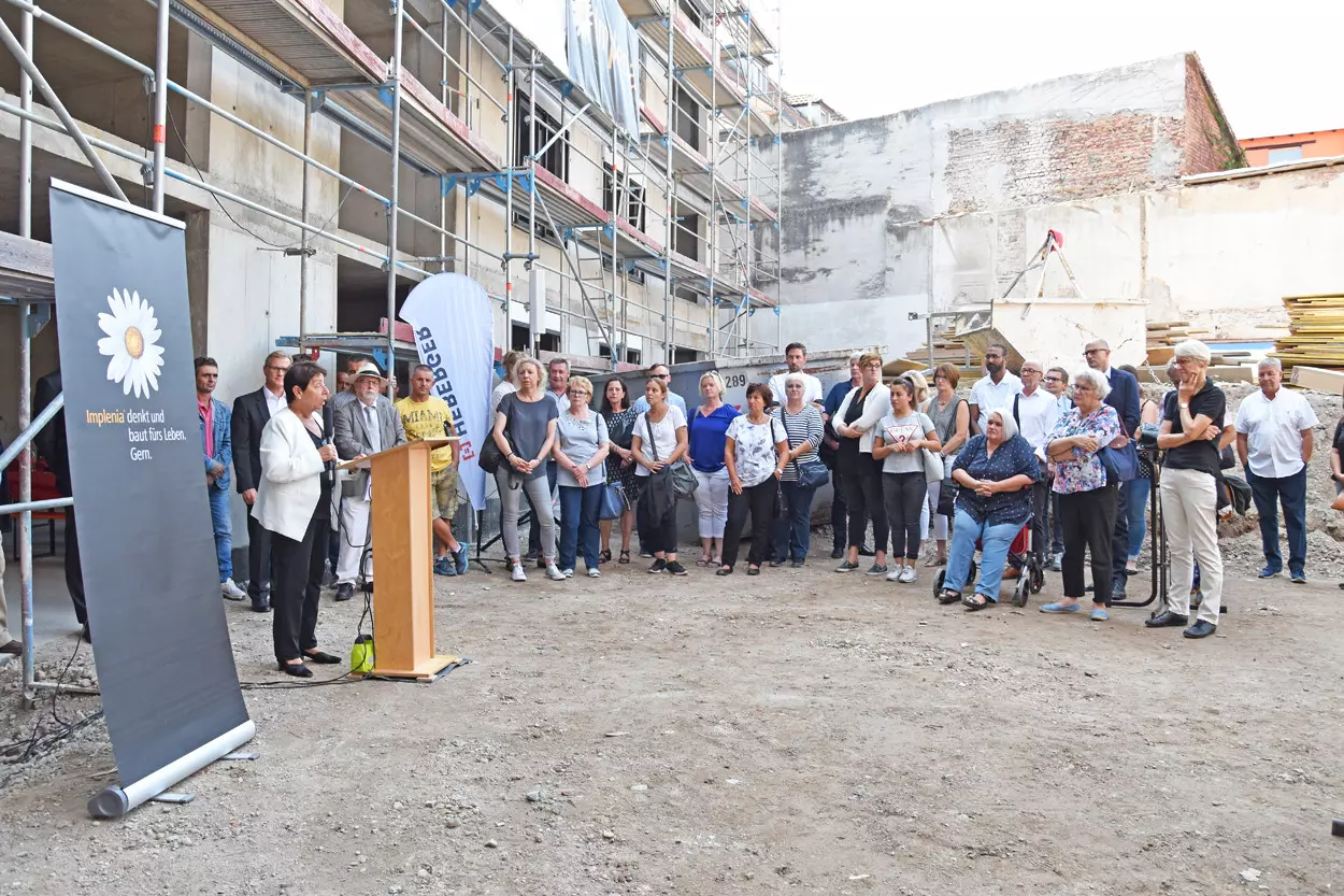 Eine Frau steht an einem Rednerpult auf einer Baustelle, viele Menschen stehen in einem Halbkreis daneben.