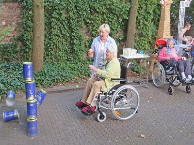 Eine Seniorin sitzt im Rollstuhl und wirft einen Ball auf eine Dosenpyramide.