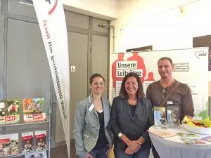 Zwei Frauen und ein Mann stehen vor einem AWO Roll-up und lächeln.