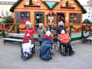 Drei Frauen sitzen im Rollstuh. Hinter ihnen stehen drei Frauen und man sieht dahinter eine Weihnachtsmarkt-Holzhütte.