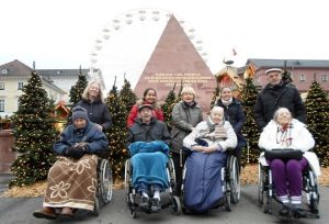 Frauen und Männer sitzen in Rollstühlen vor der Karlsruher Pyramide auf dem Marktplatz, dahinter ist das Glücksrad vor dem Schloss zu sehen.