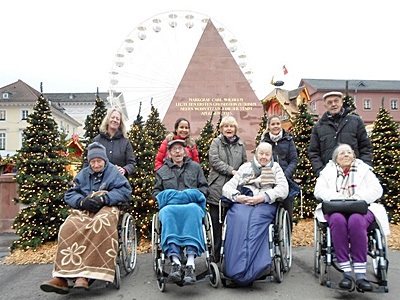 Frauen und Männer sitzen in Rollstühlen vor der Karlsruher Pyramide auf dem Marktplatz, dahinter ist das Glücksrad vor dem Schloss zu sehen.