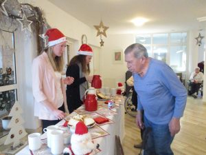 Ein Mann steht vor einem Tisch und holt sich Glühwein von zwei Frauen mit Weihnachtsmann-Mütze.