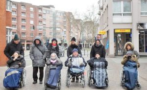 Menschen stehen und sitzen im Rollstuhl in der Stadt und schauen in die Kamera.