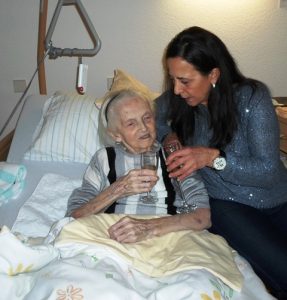 Eine Frau mit dunklen, langen Haaren, sitzt neben einer Seniorin auf dem PFlegebett und prostet ihr zu.