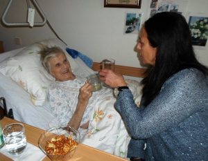 Eine Frau mit dunklen, langen Haaren, sitzt neben einer Seniorin auf dem PFlegebett und prostet ihr zu.