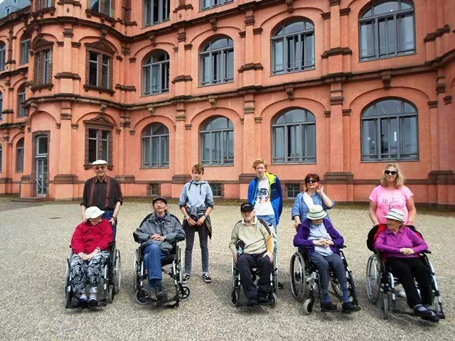 Senioren sitzen im Rollstuhl vor dem Schloss Gottesaue mit zwei jungen Männern.
