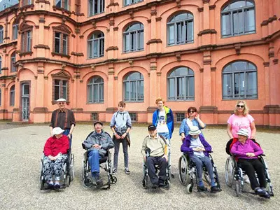 Senioren sitzen im Rollstuhl vor dem Schloss Gottesaue mit zwei jungen Männern.