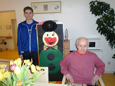 Ein junger Mann und eine Seniorin stehen neben einer Clownsfigur aus Pappe.