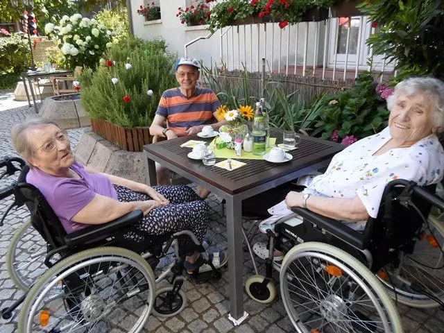 Drei Seniorinnen sitzen gemeinsam an einem Tisch und schauen fröhlich in die Kamera