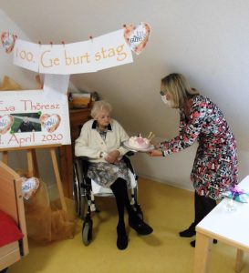 Eine Altenpflegerin überreicht einer Seniorin, die im Rollstuhl sitzt, eine Torte zu Ihrem 100. Geburtstag.