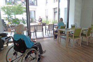 Seniorinnen sitzen in Rollstühlen in einem Haus und schauen in einen Innehofe.