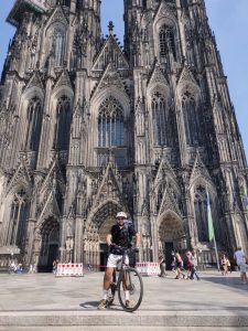 Ein Manns steht mit seinem Fahrrad vor dem Köln.