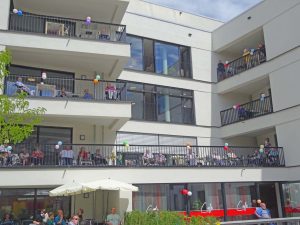 Senior*innen sitzen auf ihren Balkonen und winken.