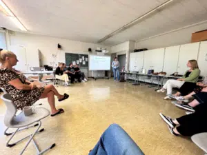Erwachsene sitzen im Stuhlkreis in einem Klassenraum, eine Person hält vorne eine Präsentation.