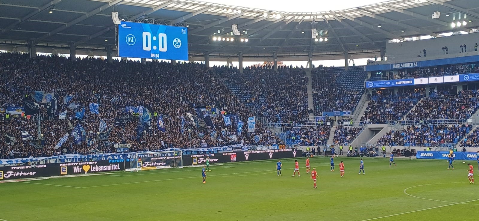 Das voll besetzte Wildparkstadion in Karlsruhe. Auf der Anzeigetafel steht 0:0 zwischen dem Karlsruher SC und dem 1. FC Kaiserslautern. Die Fankurve des KSC ist in Blau gekleidet, viele schwenken Fahnen. Auf dem Spielfeld stehen Spieler beider Mannschaften, in Blau und in Rot.