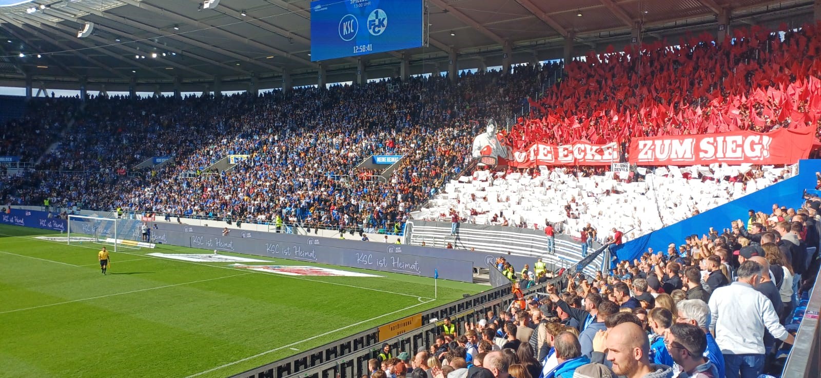 Blick in das Stadion mit zwei Fanblöcken: Links die blau-weiße KSC-Fankurve, rechts die rot-weiße Kurve der Gäste mit einer großen Choreografie und dem Transparent „In der Ecke zum Sieg!“. Die Sonne scheint auf das Spielfeld.