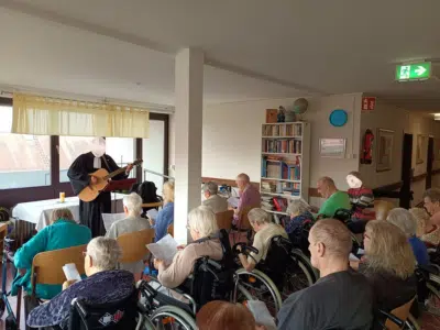 Pfarrer mit Gitarre leitet einen Gottesdienst im Gemeinschaftsraum des Haus am Blumentor. Viele ältere Bewohnerinnen und Bewohner, einige im Rollstuhl, sitzen in Reihen und singen mit. Die Atmosphäre ist ruhig und aufmerksam.