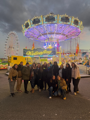 Eine Gruppe von etwa fünfzehn Menschen steht dicht beisammen vor einem beleuchteten Kettenkarussell auf einem Jahrmarkt. Hinter ihnen leuchten bunte Lichter, ein großes Riesenrad und das Karussell mit der Aufschrift „Wellenflug“. Die Personen tragen warme Kleidung, einige lächeln in die Kamera, und der Himmel ist dunkelgrau mit Abendstimmung.