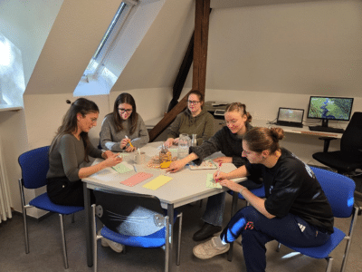 Fünf junge Erwachsene sitzen gemeinsam an einem Tisch in einem hellen Raum mit Dachschräge. Sie arbeiten konzentriert an farbigen Karten, lächeln und tauschen sich aus. Auf dem Tisch liegen Schreibmaterialien und Wasserflaschen, im Hintergrund stehen zwei Computerbildschirme.