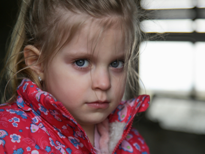 Das Bild zeigt ein kleines Mädchen mit blondem Haar, das zu einem halben Zopf gebunden ist. Sie trägt eine rote Jacke mit buntem Blumenmuster und blickt direkt in die Kamera. Ihr Gesichtsausdruck wirkt ernst. Der Hintergrund ist unscharf und zeigt einen Innenraum mit wenig Licht, sodass der Fokus auf dem Gesicht des Kindes liegt.