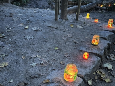 Im Außenbereich einer Kita stehen selbstgebastelte, leuchtende Windlichter aus bemalten Gläsern am Rand eines sandigen Weges. Herbstblätter liegen auf dem Boden, der beleuchtete Weg führt durch den Garten.