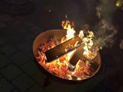 Nahaufnahme einer Feuerschale mit brennenden Holzscheiten und glühender Kohle. Flammen und Rauch steigen in die Dunkelheit auf.