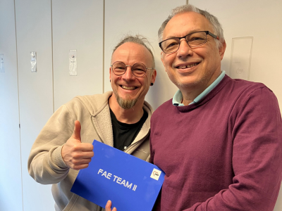 Zwei erwachsene Männer stehen nebeneinander vor einer hellen Wand. Beide lächeln in die Kamera. Der Mann links trägt eine helle Jacke, zeigt mit der rechten Hand den Daumen nach oben und hält einen blauen Umschlag mit der Aufschrift „FAE TEAM II“. Der Mann rechts trägt einen bordeauxfarbenen Pullover und hält den Umschlag gemeinsam mit ihm.