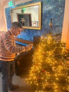 Ein Mann steht vor einem geschmückten Weihnachtsbaum und bringt eine Lichterkette an. Der Baum ist mit goldenen Kugeln und warmem Lichterglanz dekoriert.