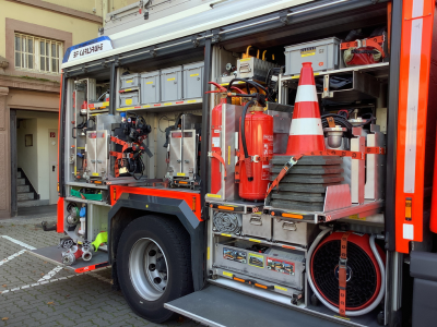 Geöffnete Seitenfächer eines Feuerwehrfahrzeugs mit detailliert sichtbarer Ausrüstung. Zu sehen sind Feuerlöscher, Verkehrsleitkegel, Werkzeuge, Schläuche und Materialboxen.