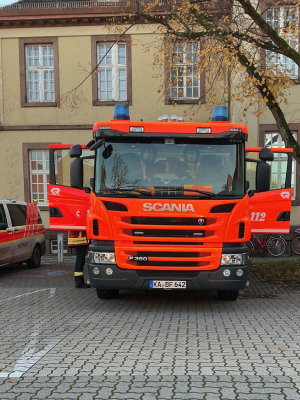 Frontansicht eines Feuerwehrfahrzeugs der Marke Scania auf einem gepflasterten Hof. Beide Türen stehen offen und Einsatzkräfte sind im Inneren erkennbar.