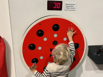 Ein Kind steht vor einer runden, roten Reaktionswand mit schwarzen Druckfeldern. Es streckt die Hand aus, um eines der Felder zu berühren. Über der Wand ist eine digitale Anzeige mit einer Zahl angebracht. Die Szene zeigt spielerisches Lernen und Bewegung.