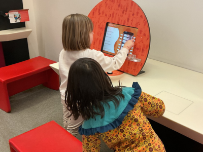 Zwei Kinder stehen an einem interaktiven Bildschirm und berühren gemeinsam die Anzeige. Sie lehnen sich leicht nach vorne und konzentrieren sich auf den Inhalt. Der Bildschirm ist in eine kindgerechte Station integriert, daneben stehen rote Sitzmöbel.