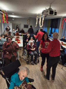 Bewohner*innen sitzen und stehen in einem Gemeinschaftsraum, einige nutzen Rollstühle. Der Raum ist weihnachtlich geschmückt, mehrere Personen bewegen sich zur Musik.