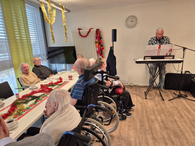 Ein Musiker spielt Keyboard vor sitzenden Bewohner*innen, darunter mehrere Personen im Rollstuhl. Die Tische sind mit weihnachtlicher Dekoration geschmückt, im Hintergrund hängen Girlanden.