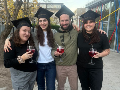 Vier junge Erwachsene stehen draußen nebeneinander, tragen schwarze Doktorhüte und halten Gläser mit rotem Getränk in der Hand. Sie lächeln in die Kamera und haben die Arme umeinander gelegt. Im Hintergrund stehen Bäume und ein Gebäude mit bunten Streben.