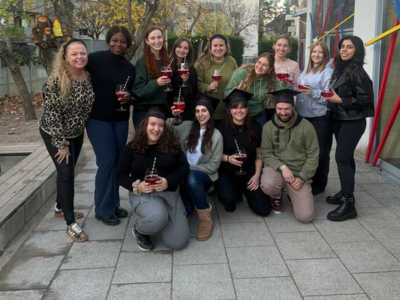 Eine größere Gruppe junger Menschen steht und kniet vor einem Gebäude. Einige tragen schwarze Doktorhüte, alle halten Gläser mit rotem Getränk. Die Stimmung wirkt fröhlich und ausgelassen. Rechts sind bunte Streben an der Fassade zu sehen, links Bäume mit gelbem Herbstlaub.